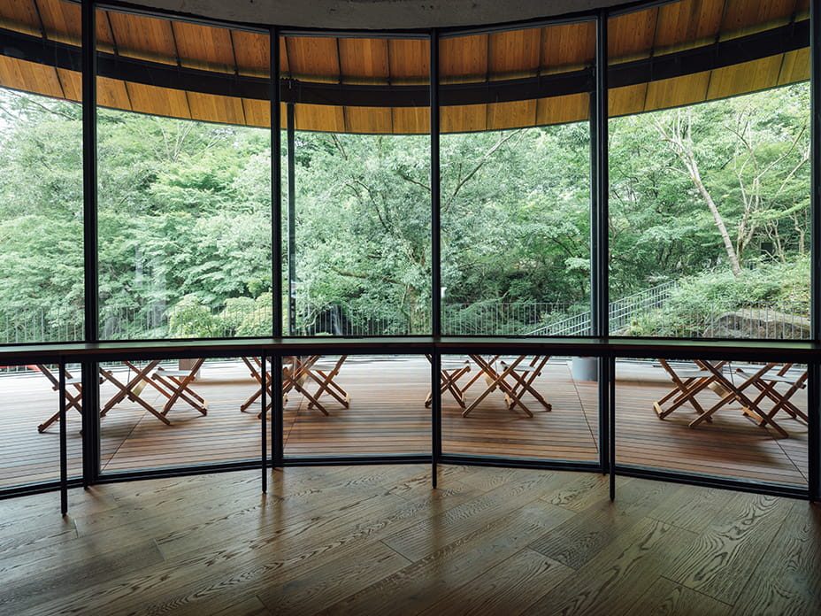 A glass-walled room with a wood floor, looking out onto the forest, with folding chairs on the deck outside