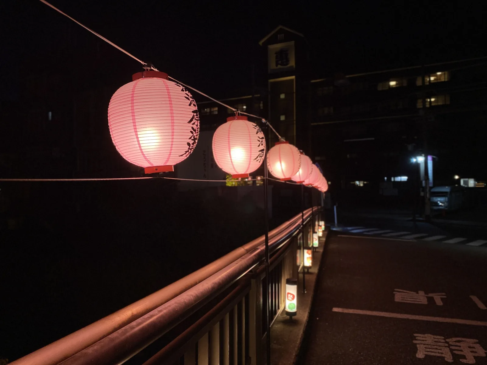 夜の橋の欄干に沿って灯るピンクの提灯。奥にホテルの建物と静かな道路が見える
