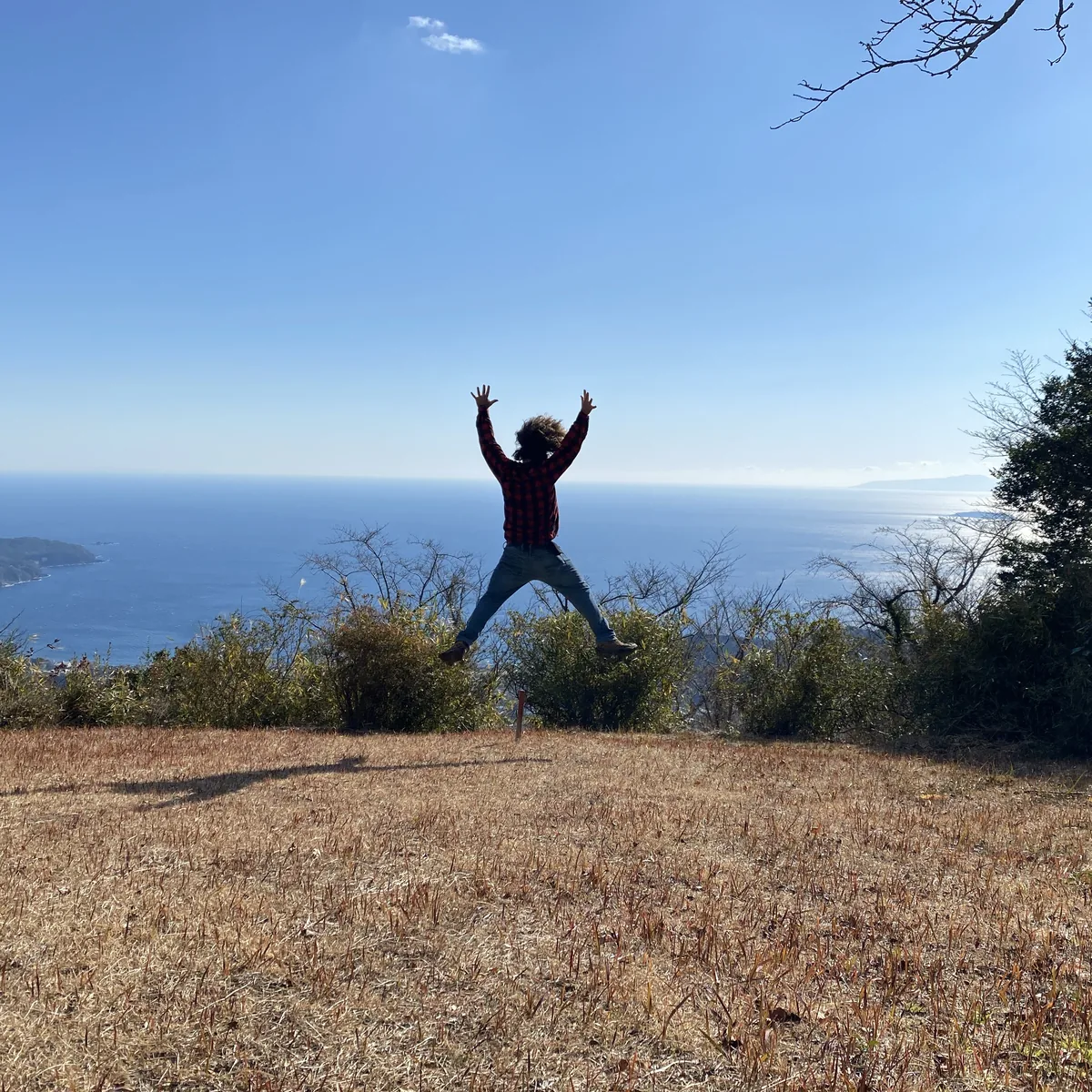 A man jumps with arms and legs spread on a grassy clearing on the Shiroyama trail, with Sagami Bay and the coast behind him under a clear sky