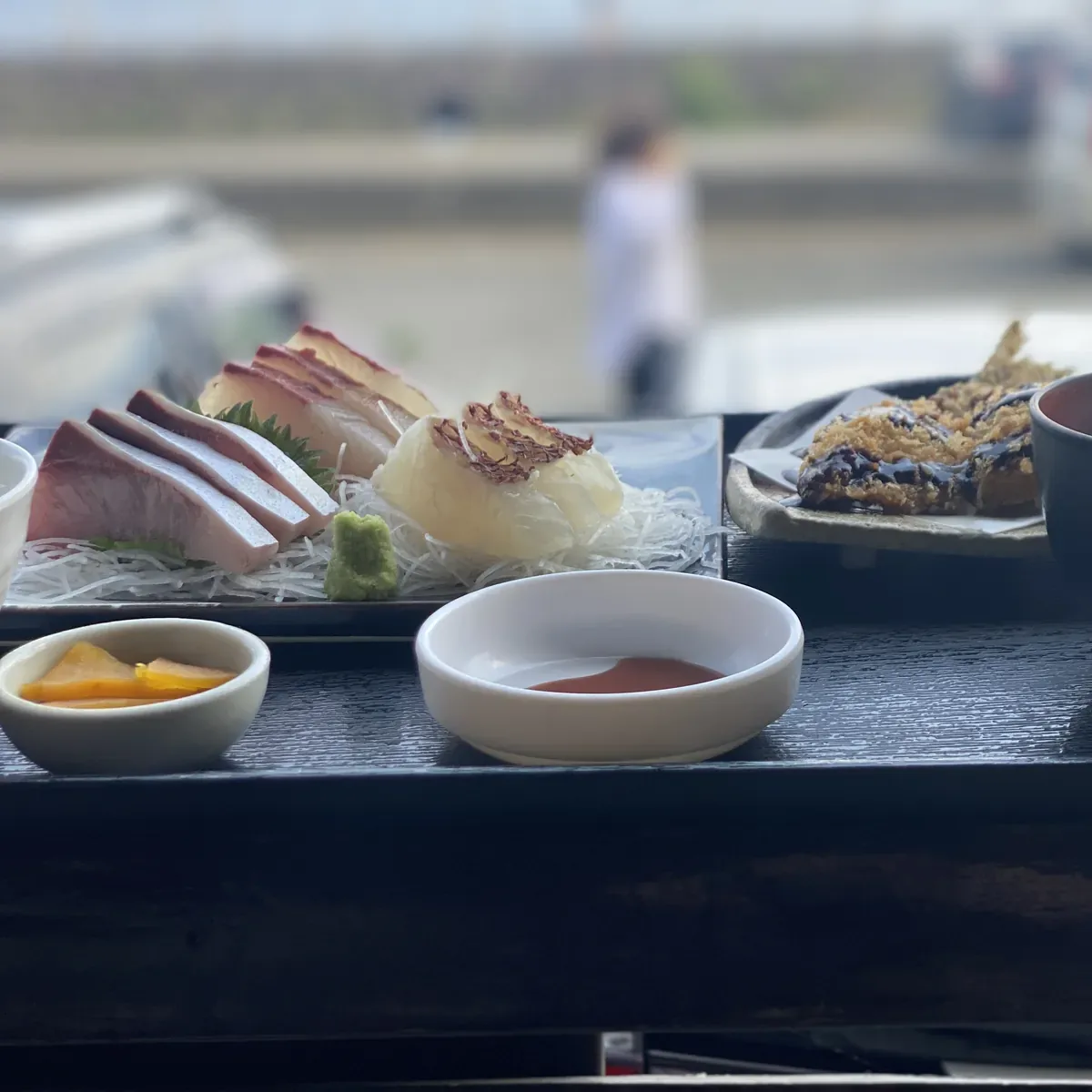 黒い盆にのった刺身定食。漬物と魚のおかず、ご飯と味噌汁の椀が端に見える